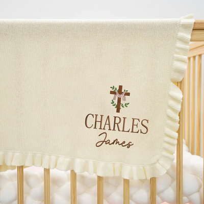 Baby blanket with embroidered cross and name 'Charles James' draped over a crib.
