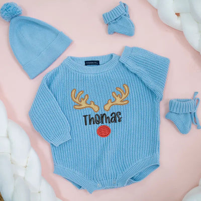 Blue baby romper with reindeer design and 'Thomas' text, accompanied by matching hat and mittens on a pink background.