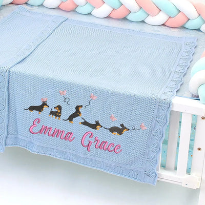 Blue personalized baby blanket with embroidered dachshunds and 'Emma Grace' text, draped over a crib.