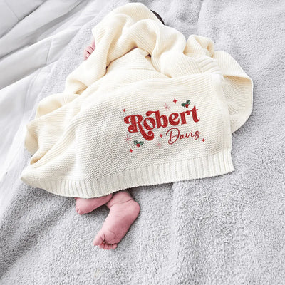 Baby wrapped in a cream personalized baby blanket with embroidered name 'Robert Davis' on a gray surface