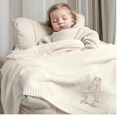 Child wrapped in a cream personalized blanket with 'Amelia' embroidered on it, lying on a white cushion.