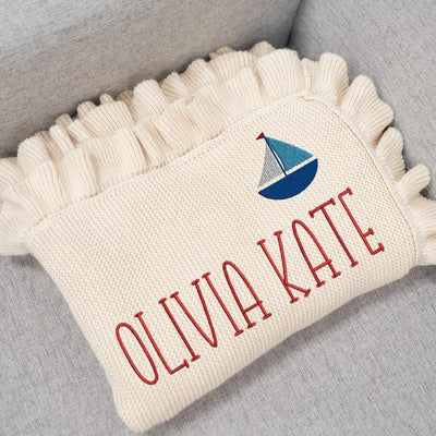 Cream personalized baby blanket with fringe, embroidered sailboat design, and 'OLIVIA KATE' text on a gray surface.